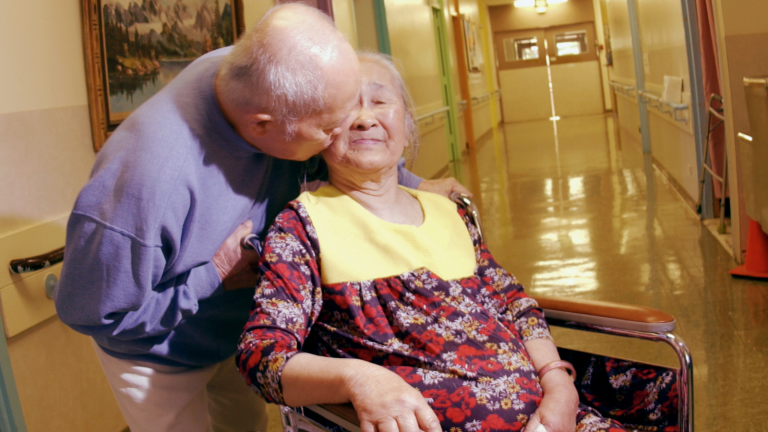 A woman in a wheelchair is kissed on the cheek by her partner