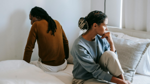 A serious couple sits on a bed facing opposite directions