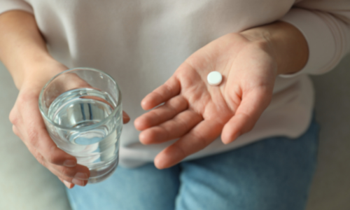 A person holding a white pill in one hand and a glass of water in another