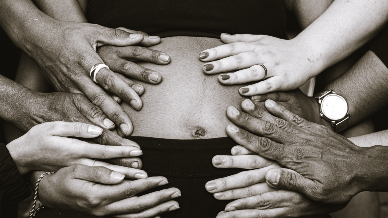 Many hands are placed on a pregnant person's belly